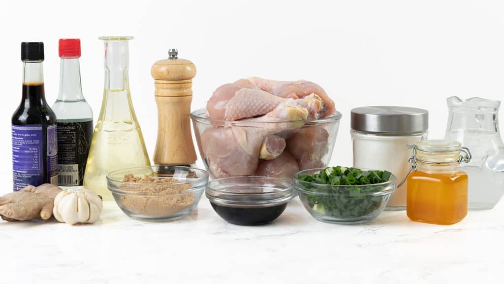 A variety of ingredients for cooking Sticky Chinese Chicken, including raw chicken, soy sauce, vinegar, pepper, sugar, ginger, garlic, chopped green onions, honey, water, and a small bottle of oil, all arranged on a white surface.