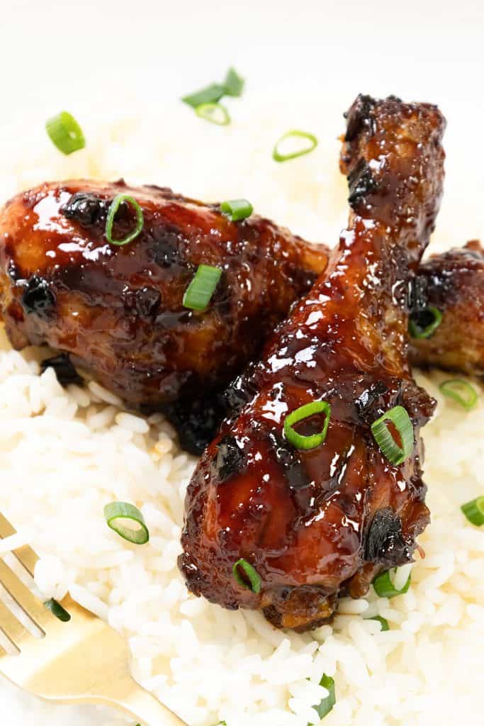 Sticky Chinese Chicken drumsticks glazed to perfection and garnished with chopped green onions, served on a bed of white rice with a gold fork beside the plate.