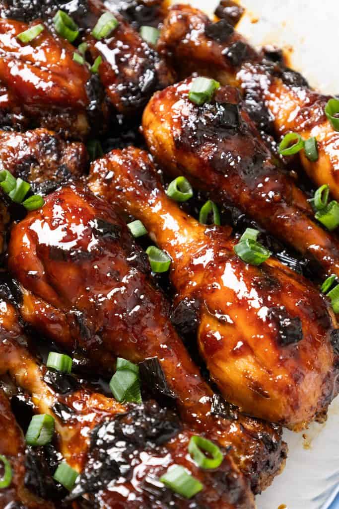 Sticky Chinese Chicken drumsticks glazed in a shiny, caramelized sauce and topped with chopped green onions, arranged closely together on a white surface.