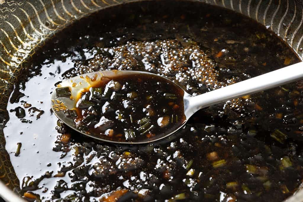 A metal spoon holds a thick, dark sauce—perfect for Sticky Chinese Chicken—with chopped green onions and garlic in a patterned bowl.