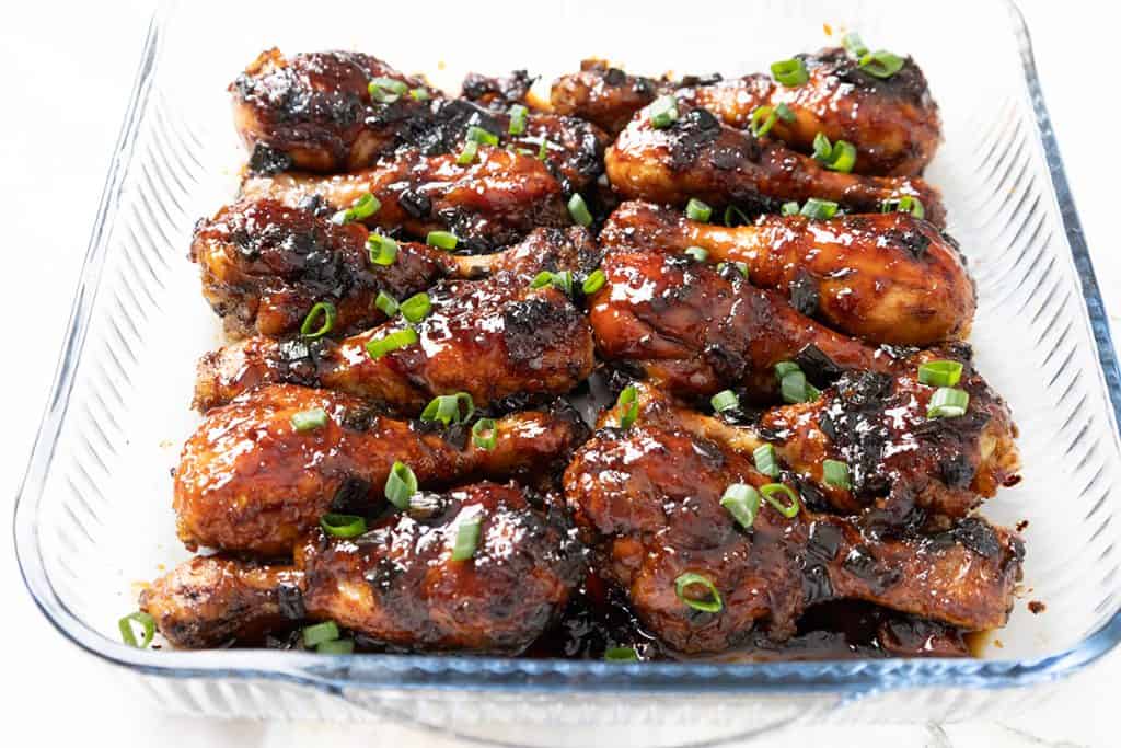 Sticky Chinese Chicken drumsticks, coated in a glossy, dark sauce, are garnished with chopped green onions and arranged neatly in a clear glass baking dish.