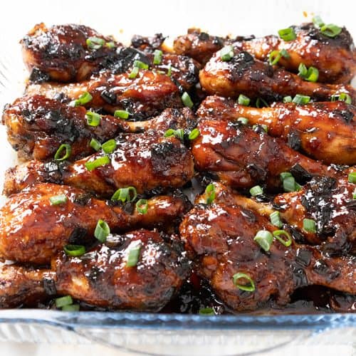 Sticky Chinese Chicken drumsticks, coated in a glossy, dark sauce, are garnished with chopped green onions and arranged neatly in a clear glass baking dish.