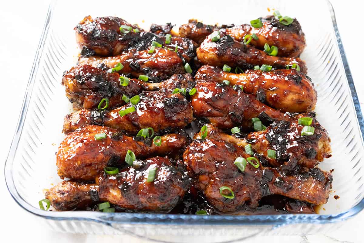 Sticky Chinese Chicken drumsticks, coated in a glossy, dark sauce, are garnished with chopped green onions and arranged neatly in a clear glass baking dish.