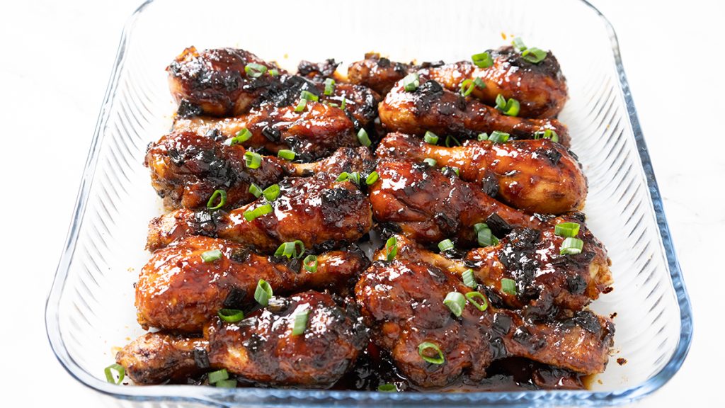 Sticky Chinese Chicken drumsticks covered in a dark, glossy sauce and garnished with chopped green onions, arranged neatly in a clear glass baking dish.