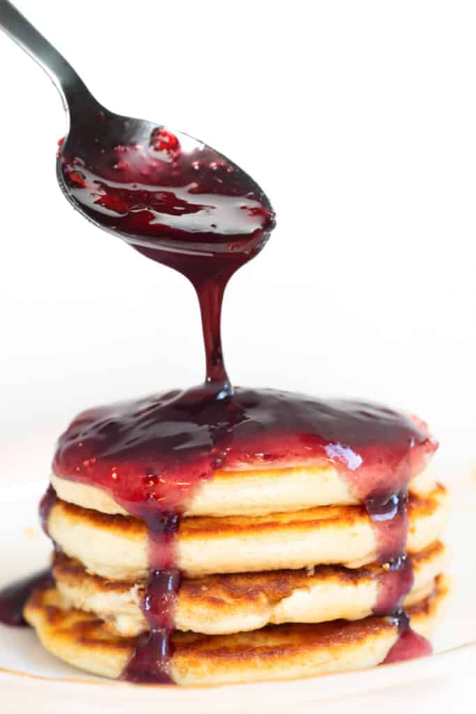 A stack of golden-brown pancakes on a plate, with a spoon drizzling dark red berry compote over the top.