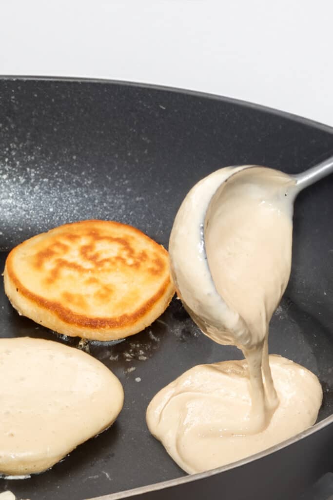 a spoon pouring scotch pancakes batter into a pan with other pancakes at different cooking stages in the pan.