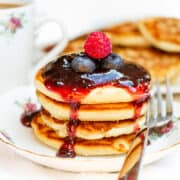A stack of Scotch pancakes topped with a glossy berry sauce and fresh berries, with a cup of tea and extra pancakes in the background, all on a white tablecloth.