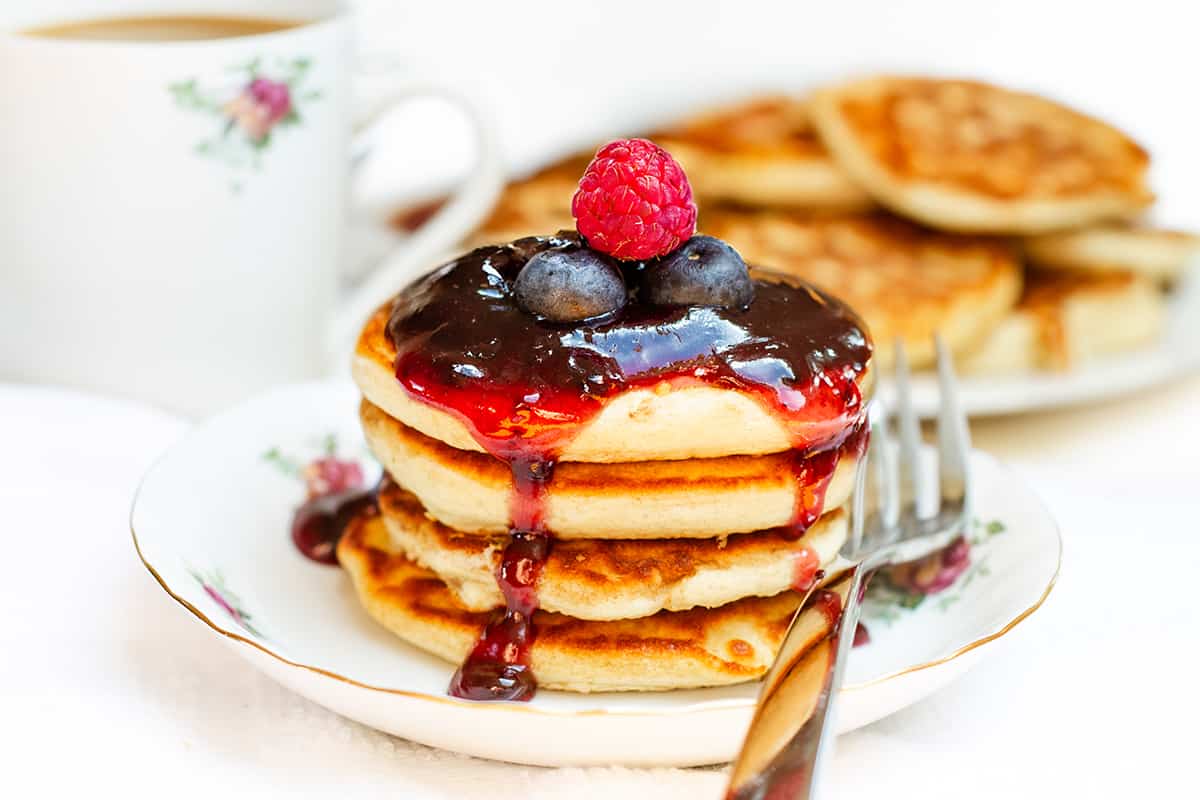 A stack of Scotch pancakes topped with a glossy berry sauce and fresh berries, with a cup of tea and extra pancakes in the background, all on a white tablecloth.