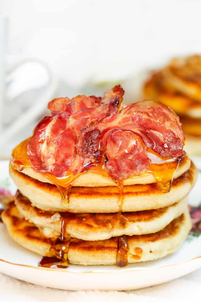 A tall stack of Scotch pancakes topped with crispy bacon strips, drizzled with golden maple syrup on a white plate.