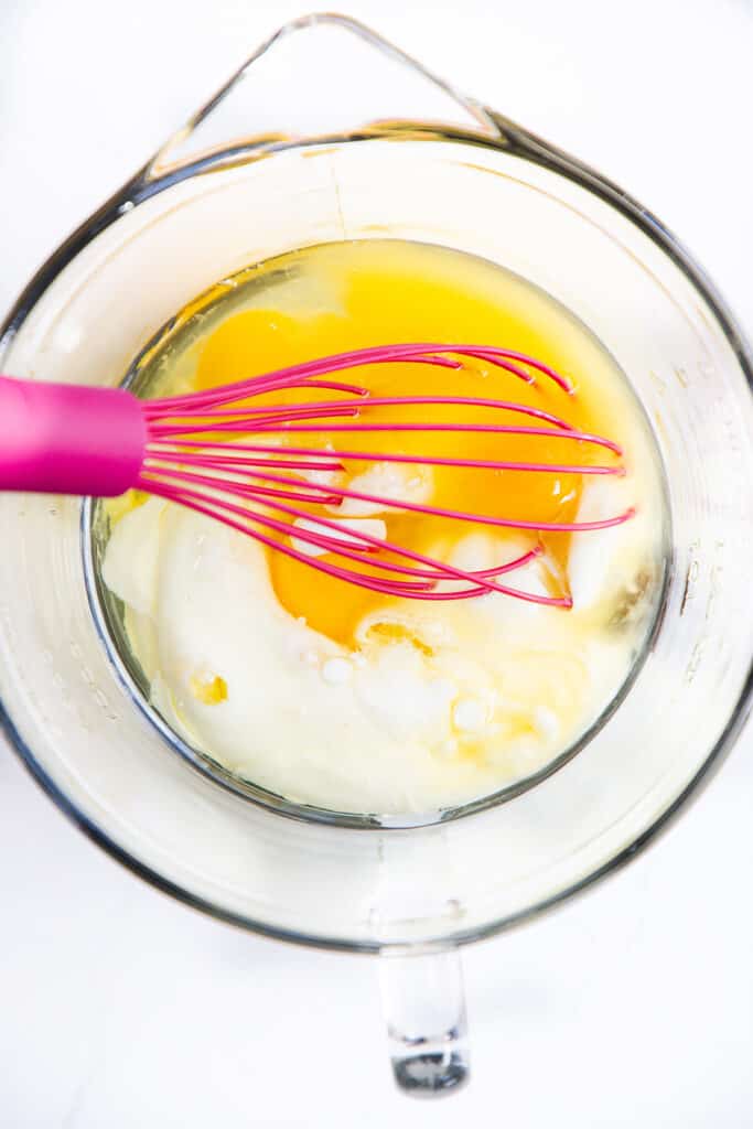 The wet ingredients for the Scotch pancakes in a glass measuring cup with a pink whisk.