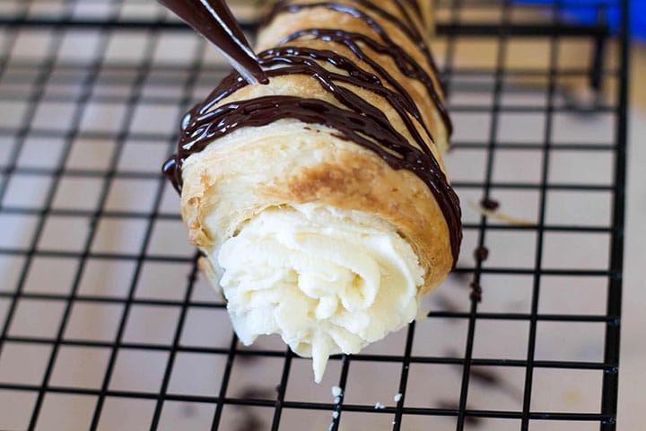 A close up of a cream horn being topped with lines of dark chocolate using a piping bag