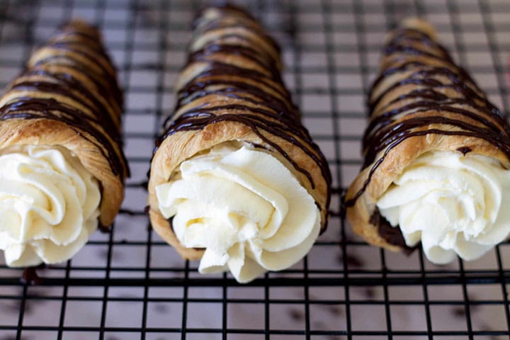 Three Dark Chocolate Cream Horns with swirled whipped cream slightly bulging out of them topped with dark chocolate drizzle.