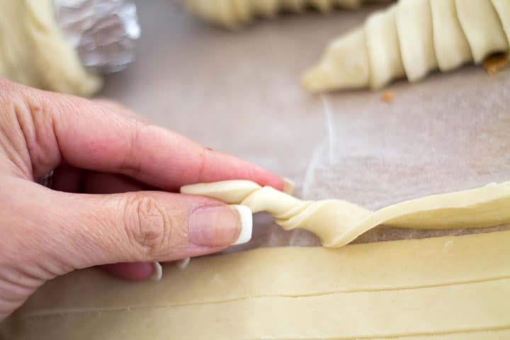 A strip of puff pastry being hand twisted surrounded by more strips and some cones covered in the puff pastry. 