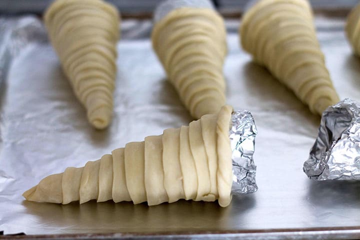 A close up of one cone wrapped with the puff pastry on a baking sheet with the rest in the background