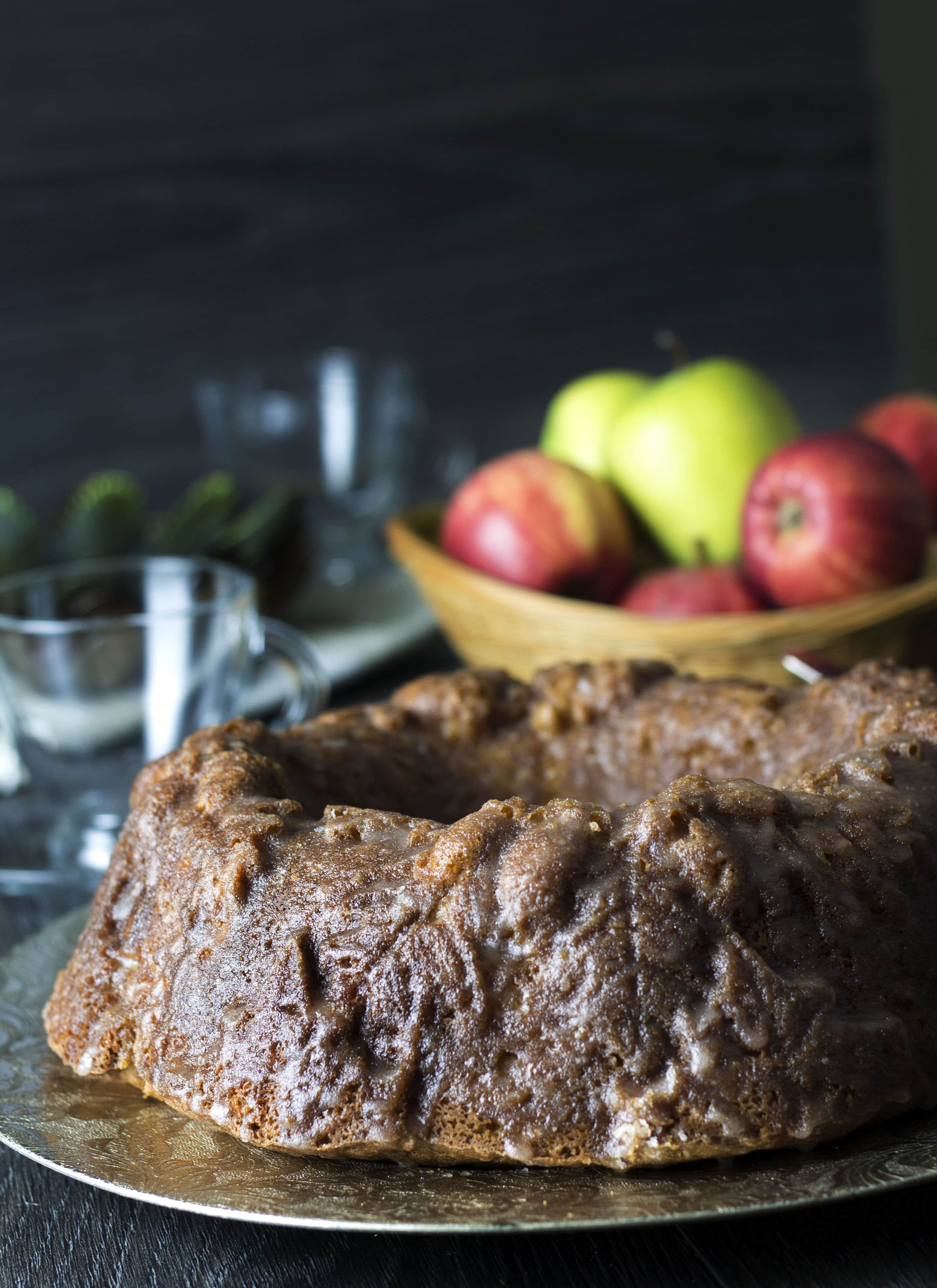 A close up of glazed apple bundt cake with a wooden bowl full of apples and drinking glasses in the background