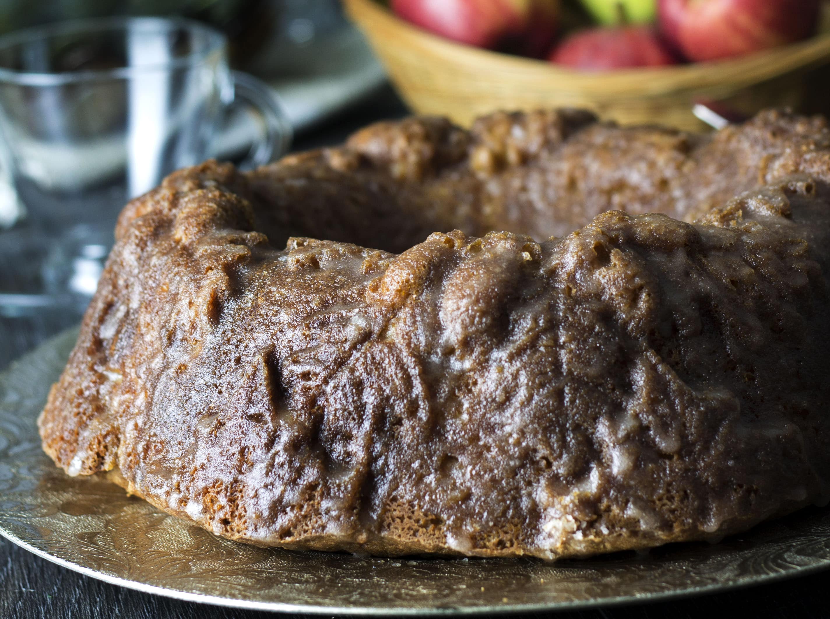 A close up of a piece of glazed apple cake with a bowl of apples in the background