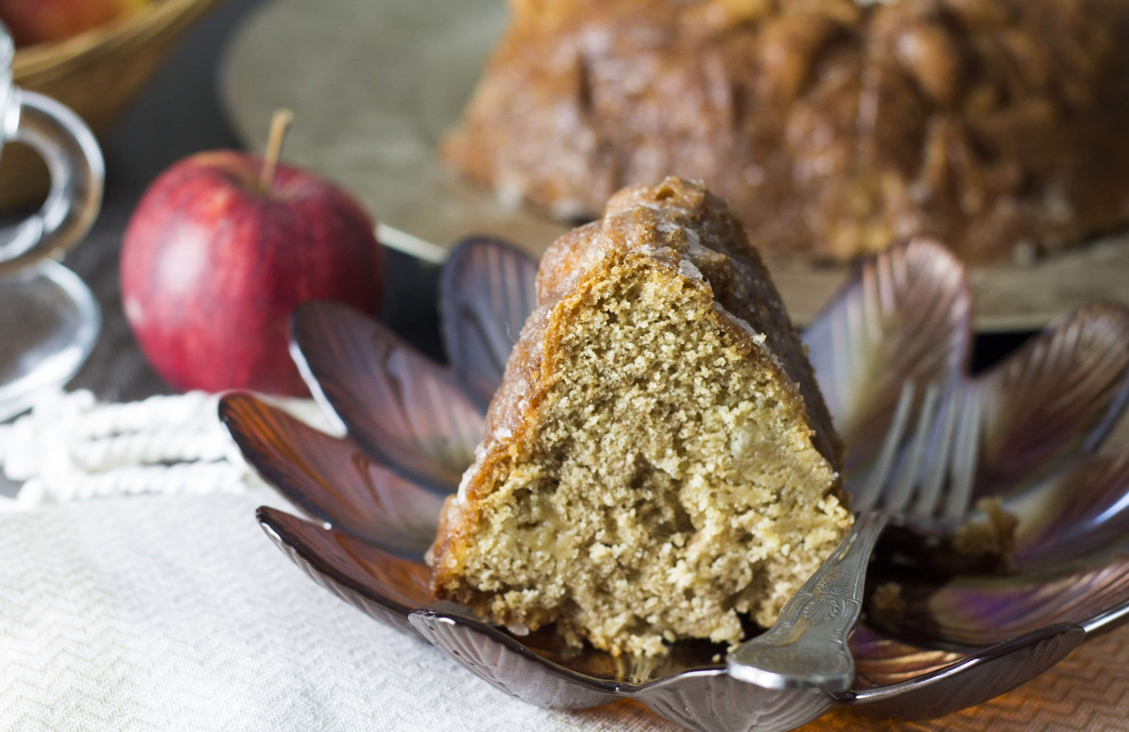 A piece of glazed apple bundt cake on a dish with an apple and the rest of the cake in the background