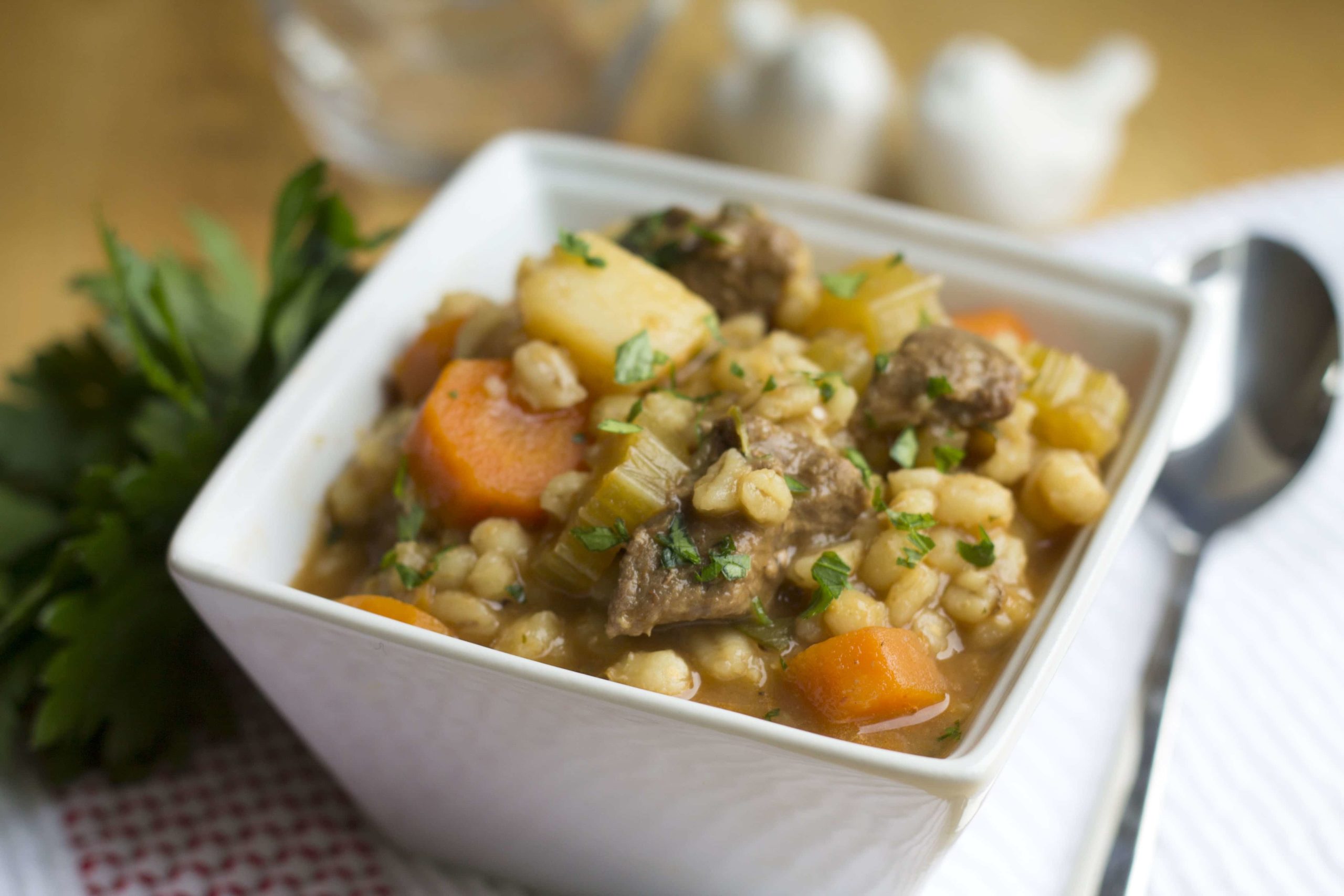 Beef & Barley Soup in a square white bowl with a spoon next to it. 