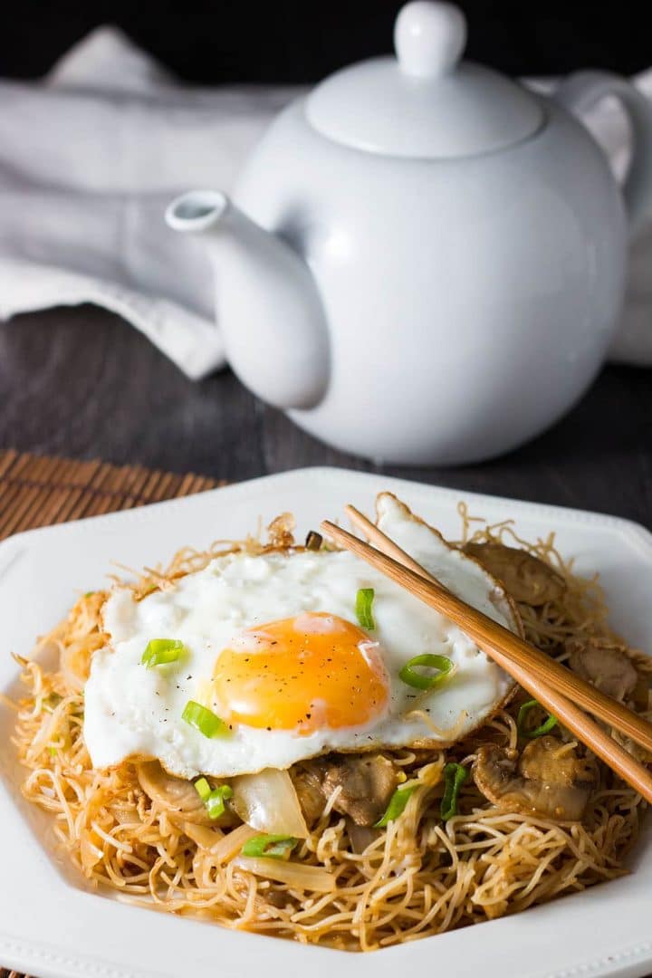 A white plate with stir fried rice noodles and vegetables topped with a fried egg and sprinkled with green onions and chopsticks laying on the plate