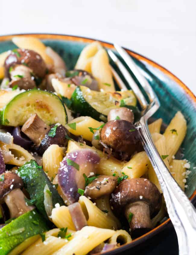 A bowl of roasted vegetable pasta with penne, sautéed zucchini, whole mushrooms, and red onions, garnished with chopped parsley and grated cheese, with a fork on the side.