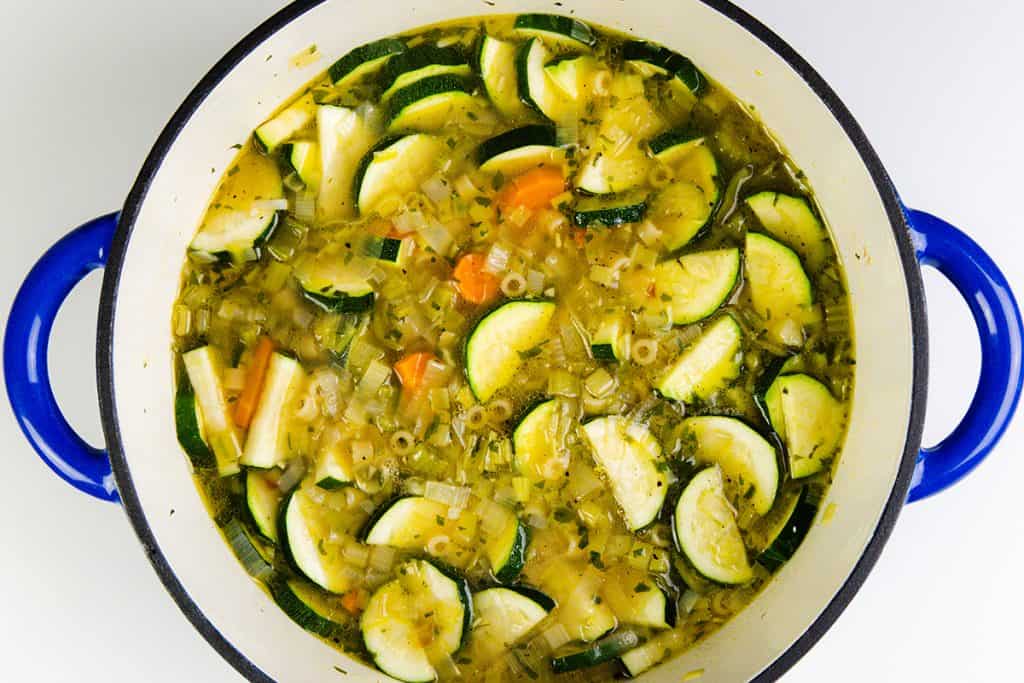A large white pot with blue handles filled with Spring Vegetable Soup, featuring sliced zucchini, carrots, celery, onions, herbs, and small pasta in a clear broth.