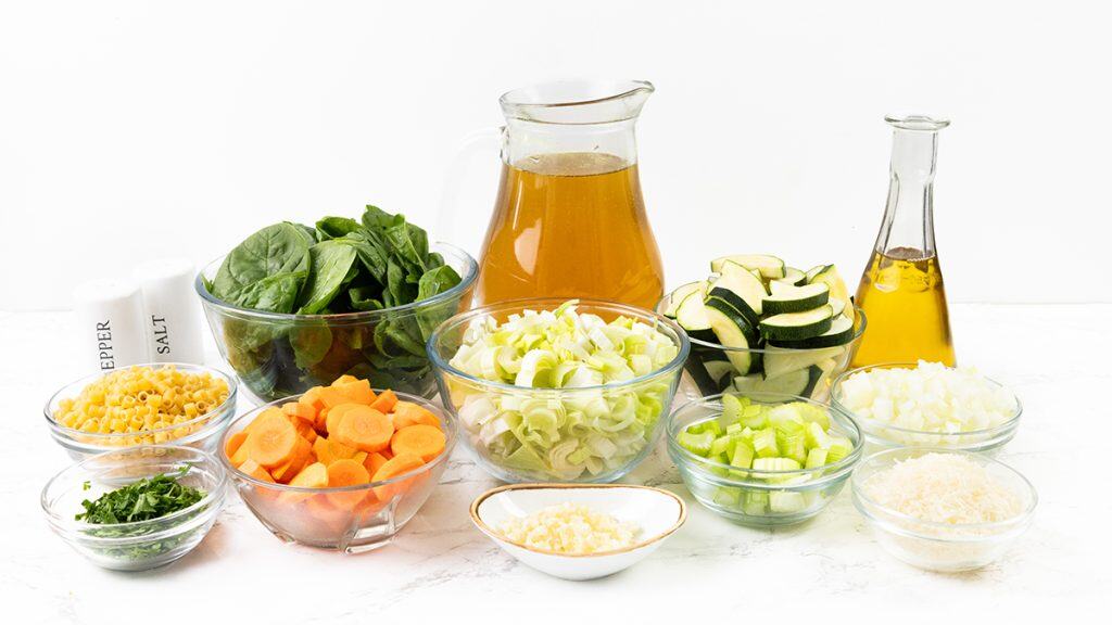 A variety of fresh Spring Vegetable Soup ingredients are arranged on a white surface, including bowls of spinach, pasta, carrots, leeks, zucchini, celery, onion, garlic, parsley, grated cheese, broth, olive oil, and seasonings.