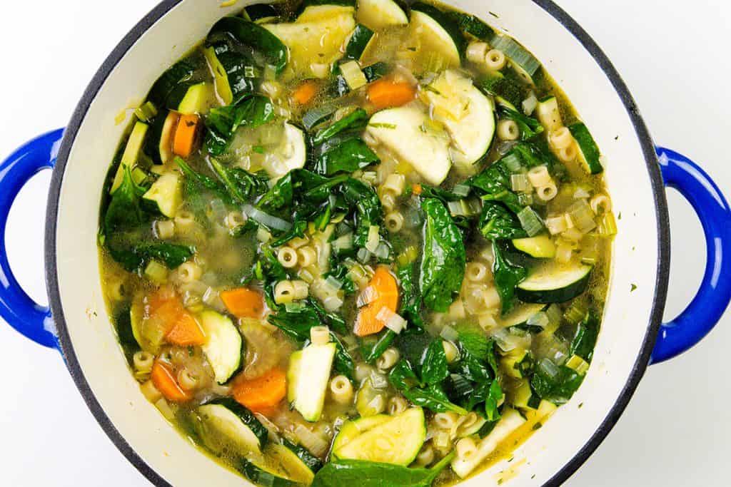 A blue-handled pot filled with Spring Vegetable Soup—spinach, carrots, zucchini, pasta, and leek pieces in a clear broth—viewed from above.