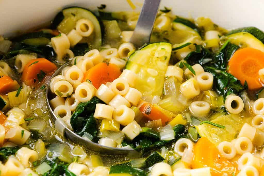 A close-up of Spring Vegetable Soup with ditalini pasta, sliced carrots, zucchini, leafy greens, and a ladle, all in a clear broth.
