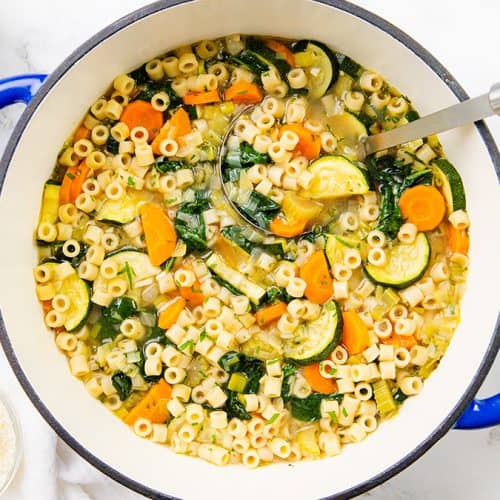 A pot of Spring Vegetable Soup with ditalini pasta, carrots, zucchini, and spinach, shown from above. A ladle rests inside the pot, next to bowls and grated Parmesan cheese on a white surface.