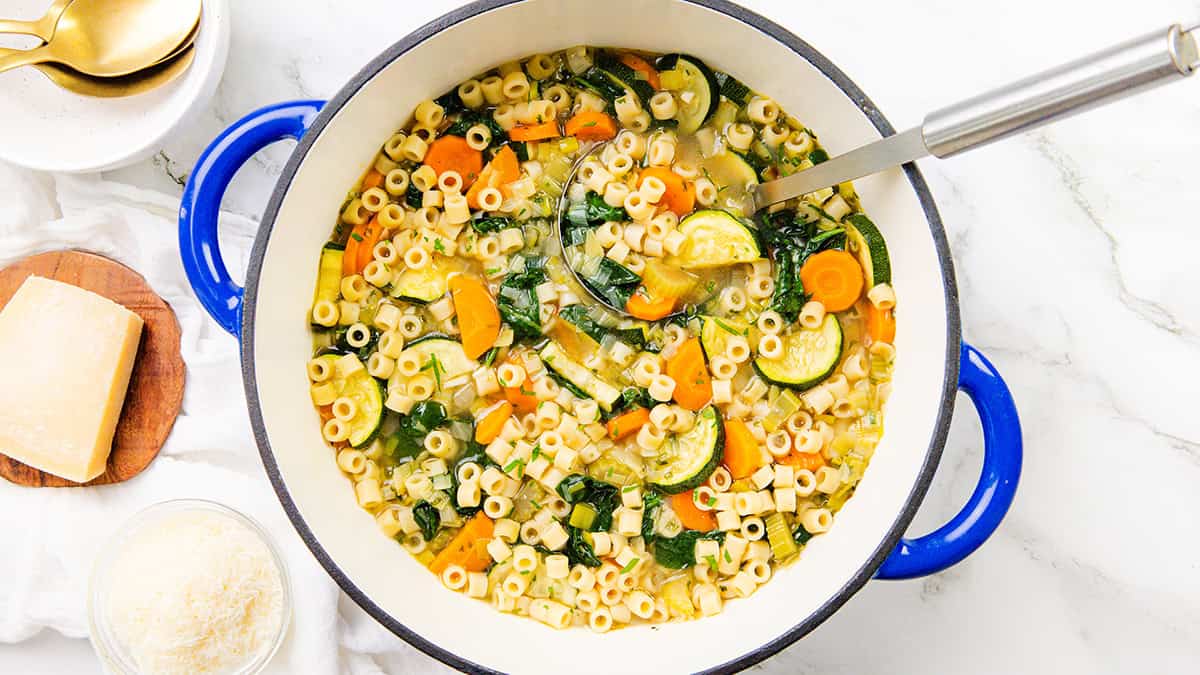 A pot of Spring Vegetable Soup with ditalini pasta, carrots, zucchini, and spinach, shown from above. A ladle rests inside the pot, next to bowls and grated Parmesan cheese on a white surface.