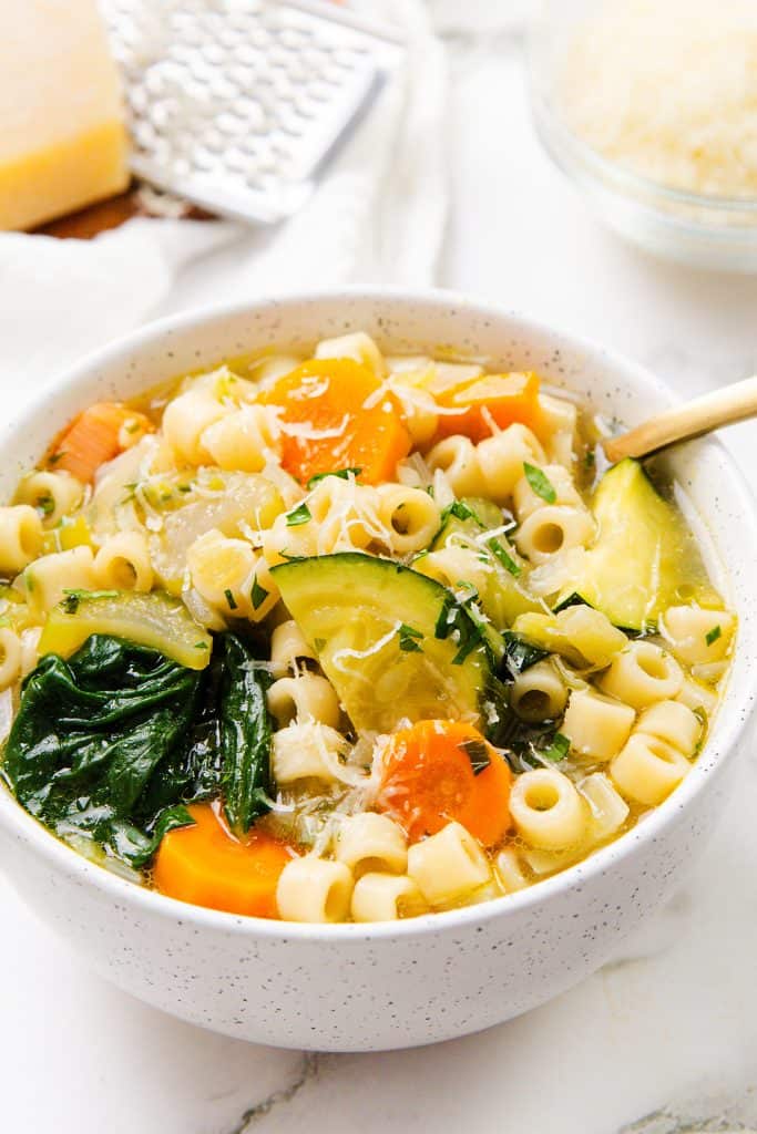 A bowl of Spring Vegetable Soup with ditalini pasta, carrots, zucchini, spinach, and grated cheese on top sits on a light surface, with a grater and cheese wedge in the background.
