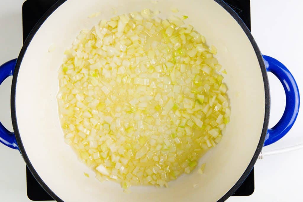 Chopped onions are being sautéed in oil in a large white pot with blue handles, placed on a black stovetop—an aromatic start for a flavorful Spring Vegetable Soup. The onions appear translucent, indicating they are being cooked.