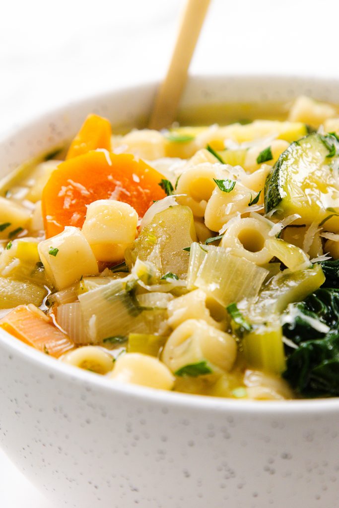 Close-up of a bowl of Spring Vegetable Soup with pasta, featuring sliced carrots, zucchini, leafy greens, and chopped herbs, with a spoon in the background.