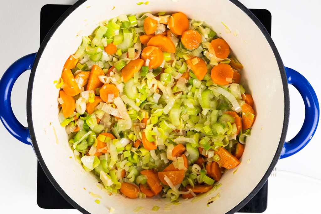 Chopped carrots, celery, and leeks are sautéing in a large white pot with blue handles on the stove—the perfect start for a fresh Spring Vegetable Soup. The vegetables appear to be in the early stages of cooking.