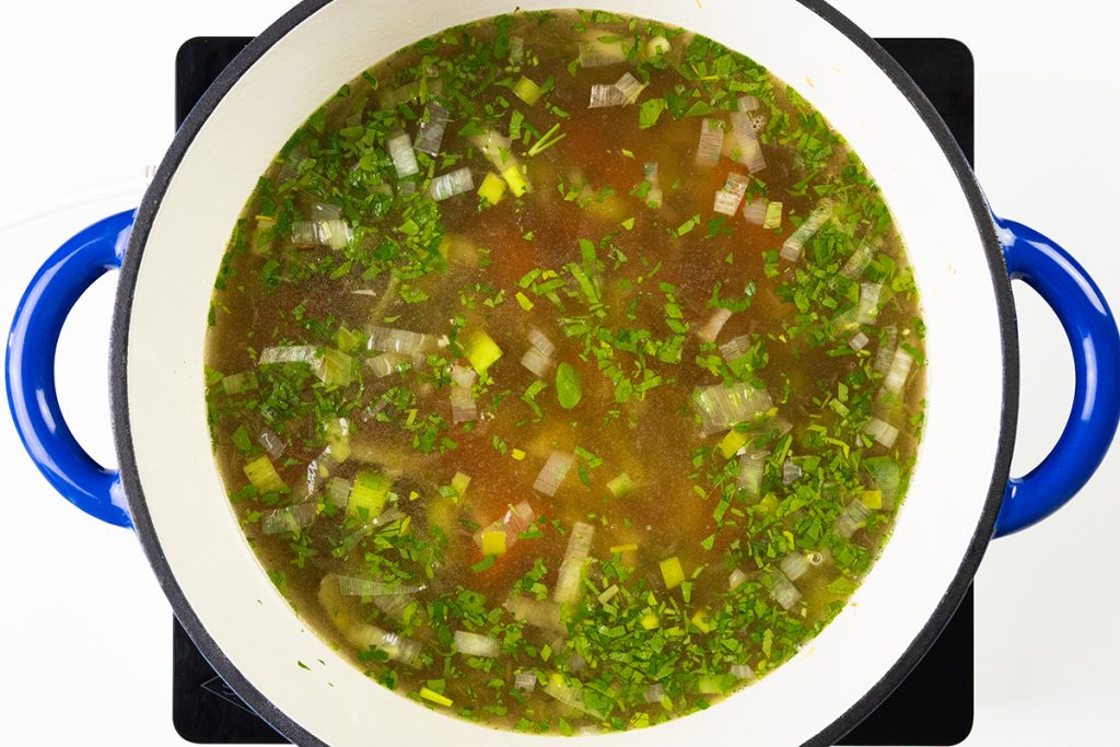 A large white pot with blue handles filled with Spring Vegetable Soup, containing chopped onions, green herbs, and broth, viewed from above. The pot is sitting on a black stovetop.