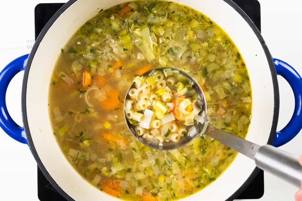 A ladle holds a serving of Spring Vegetable Soup with ditalini pasta, carrots, celery, and herbs over a pot of the same soup on a stovetop.