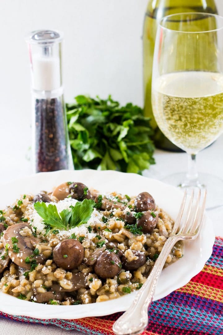 Skinny Mushroom & Barley Risotto in a white bowl and a fork next to it with a glass of white wine and salt & pepper in the background