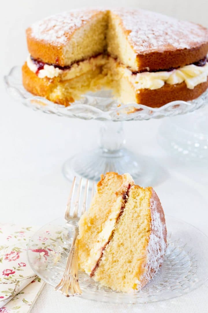 Classic Victoria Sponge Cake slice on a glass plate with fork and floral napkin with the remaining cake on a glass cake stand in the background