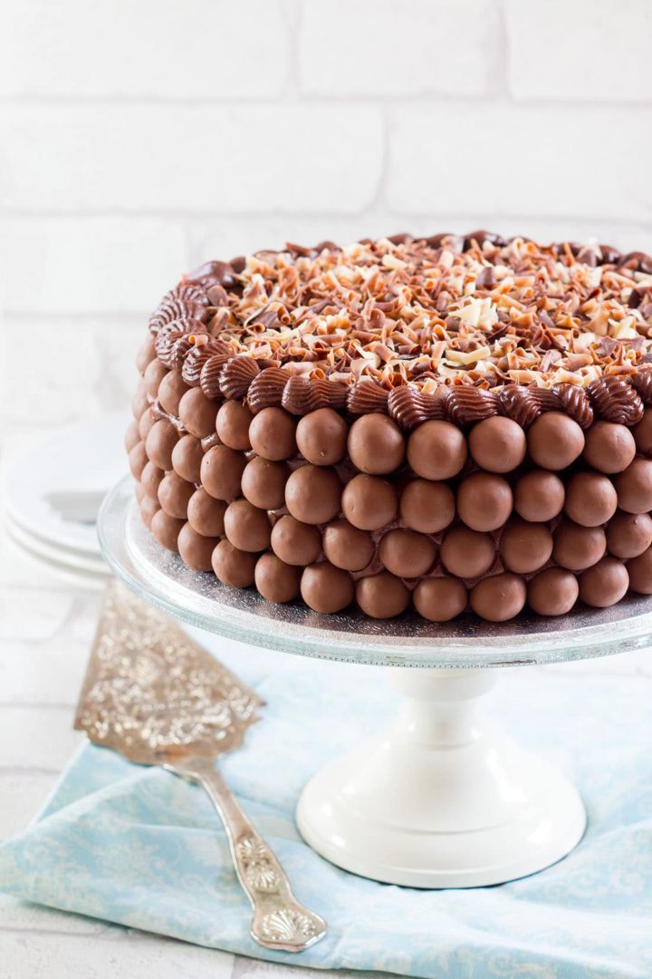 A chocolate cake covered in chocolate malt balls and chocolate flakes on a serving dish