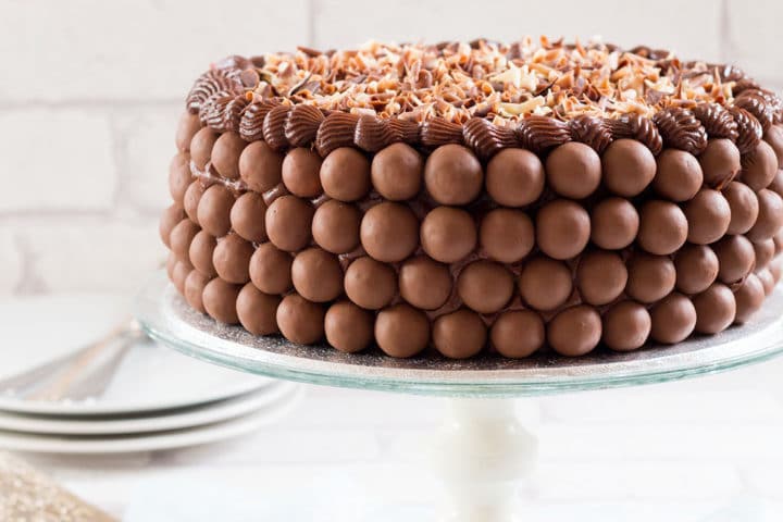 A Chocolate cake covered in malted chocolate balls and chocolate curls on a cake stand with plates and forks behind it.