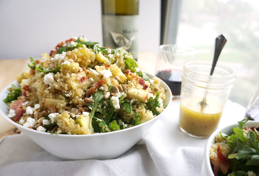 Bacon Quinoa Salad with Lemon Dijon Dressing piled high in a white bowl with a pot of dijon dressing in the background and a bottle of red wine