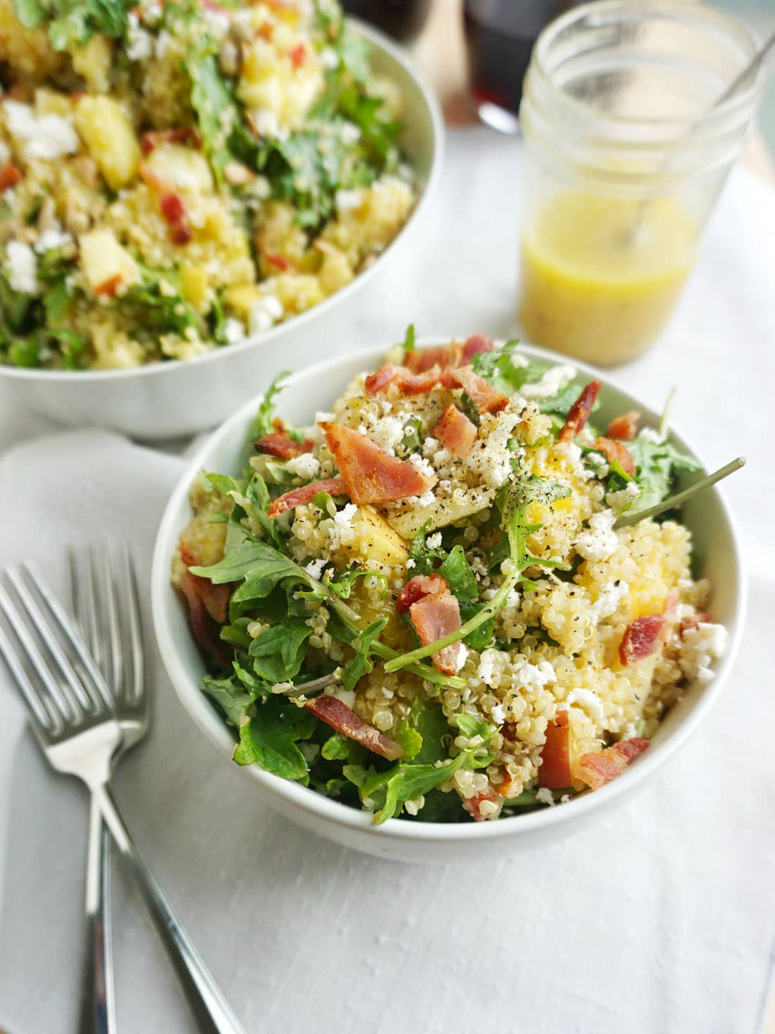 Bacon Quinoa Salad with Lemon Dijon Dressing in a bowl with two forks beside, dijon dressing in a jar with a spoon and a serving dish full of the rest of the salad in the background