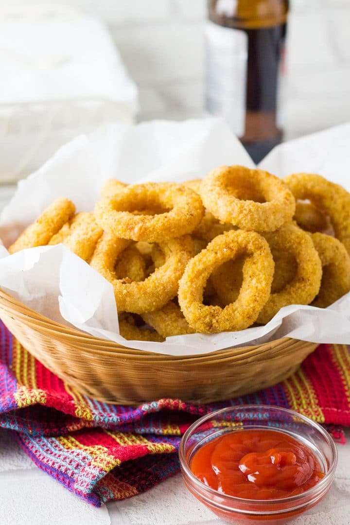 A basket full of Skinny Seasoned Baked Onion Rings with a bottle of beer in the background and a glass bowl of ketchup in front of them.