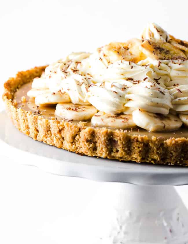 A Banoffee Pie on a cake stand showing the crust, caramel layer, banana layer, whipped cream and banana chips as decoration