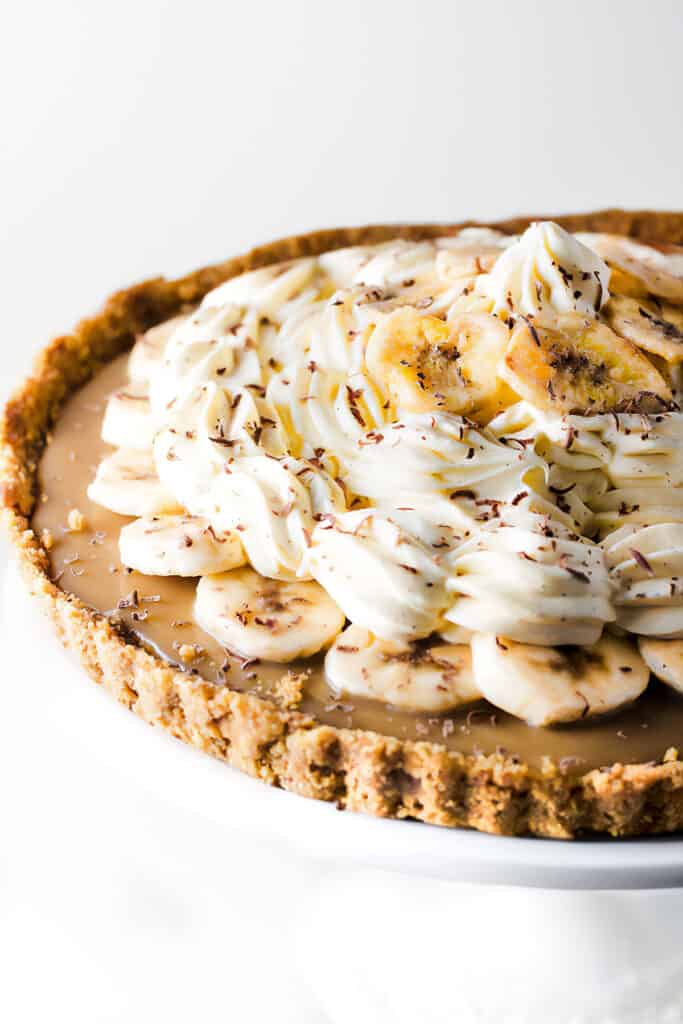 a close up image of a Banoffee Pie decorated with piped whipped cream, grated chocolate and banana chips.