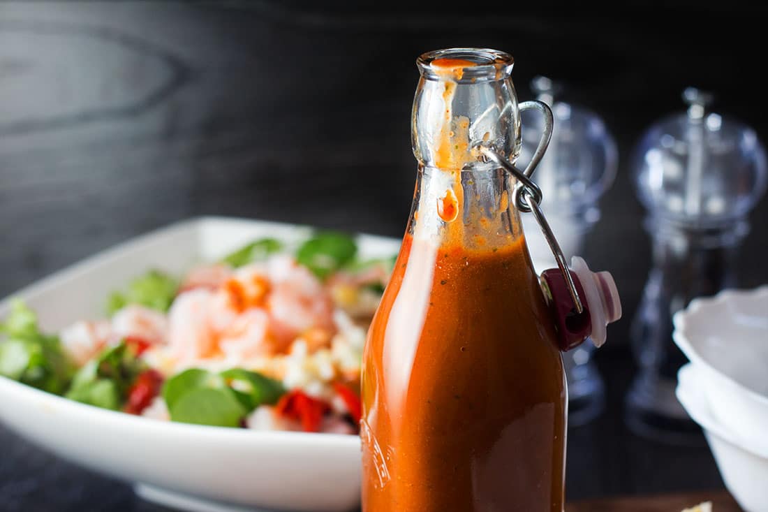 A glass bottle of Catalina style peppadew salad dressing with a bowl of food in the background