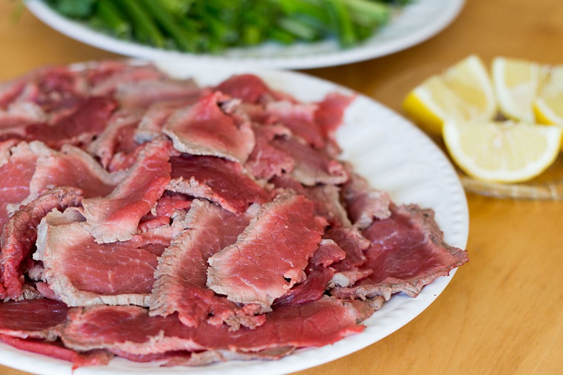 thinly sliced beef piled on a plate with a bowl of herbs and lemon wedges in the background