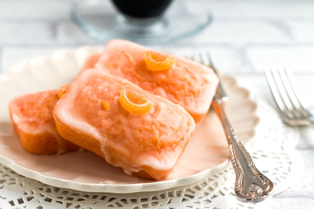 Three mini Glazed Orange Cakes on a plate with forks beside them and a cup of coffee in the background