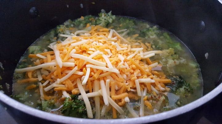 A pot with cooked broccoli florets in stock topped with a pile of shredded cheese to be stirred in.