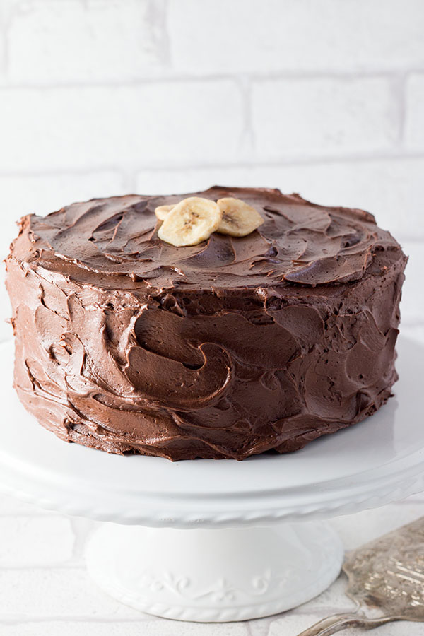 Banana Spice Cake with Chocolate Fudge Frosting on a cake stand with a background of white tiles